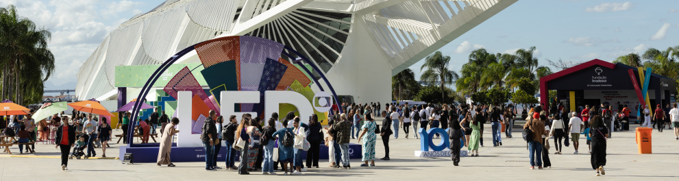 Área externa do Museu do Amanhã, no Rio de Janeiro, com o grande letreiro colorido do Festival LED em primeiro plano. Muitas pessoas circulam pelo espaço em um dia ensolarado, com céu azul e poucas nuvens.