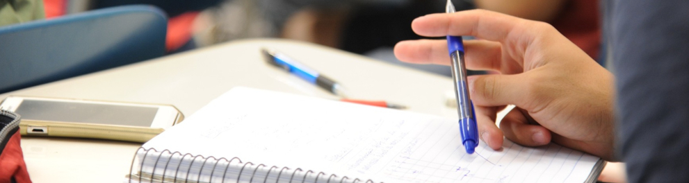 Close de um estudante usando óculos, concentrado enquanto escreve em um caderno com caneta azul; sobre a mesa há um celular e outros materiais. O fundo desfocado mostra outros alunos em sala.