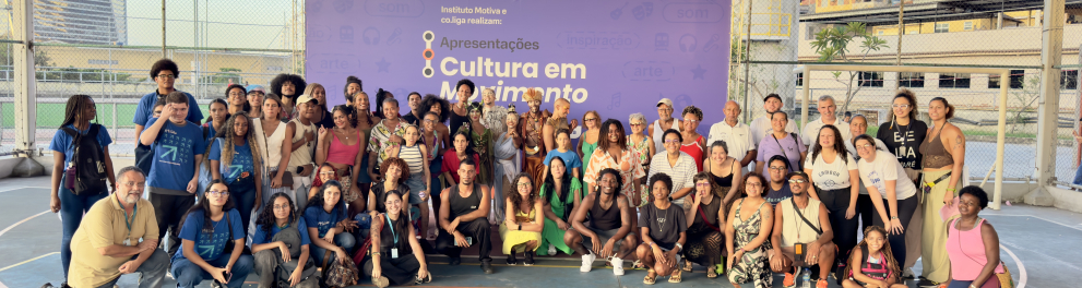 Uma grande foto de grupo reúne dezenas de pessoas em uma quadra esportiva ao ar livre, cercada por grades. Os participantes, de diferentes idades, gêneros e estilos, estão organizados em filas — alguns em pé, outros agachados — e todos voltados para a câmera, muitos sorrindo.  Ao fundo, há um painel roxo com o título “Apresentações Cultura em Movimento”, indicando um evento cultural. Também aparecem elementos urbanos ao redor, como prédios e casas em área elevada, sugerindo um contexto de comunidade.