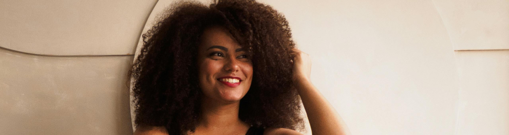 A imagem mostra uma mulher negra sentada em um sofá, com os braços apoiados no apoio do sofá e sorrindo com as mãos no cabelo e olhando para o lado.