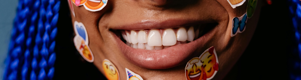 A imagem mostra uma jovem sorrindo e com diversos adesivos estampados com emojis colados em seu rosto