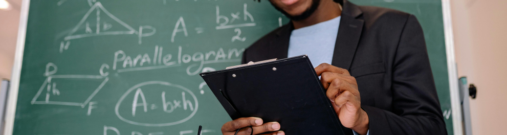 A imagem mostra um homem negro segurando uma prancheta, sorrindo e em frente a um quadro negro