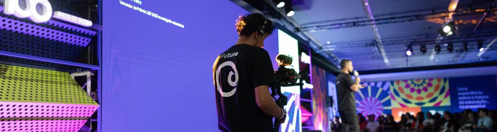 Um cinegrafista filma uma apresentação em um palco durante um evento. Ele aparece em primeiro plano, vestido com camiseta preta e usando equipamentos de gravação presos ao corpo. À frente dele, um homem segura um microfone e fala para uma plateia, embora esteja desfocado ao fundo.  O palco é grande e moderno, iluminado por luzes em tons de azul, roxo e rosa