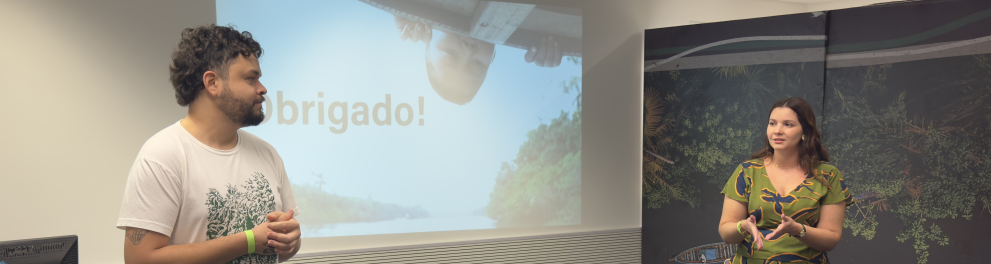 Em uma sala de apresentação, as mesmas duas pessoas aparecem em pé diante de um público. À esquerda, o homem de camiseta branca e calça bege observa a colega enquanto mantém as mãos unidas à frente do corpo. À direita, a mulher de macacão estampado fala gesticulando com as mãos. Atrás deles, um telão projeta a palavra “Obrigado!” sobre uma imagem de rio cercado por vegetação. Ao lado direito há um painel decorativo com fotografia aérea de floresta amazônica. 