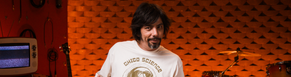 Homem de cabelos médios e escuros posa sentado em um estúdio musical iluminado por luzes alaranjadas. Ele usa camiseta clara estampada com a frase “Chico Science” e aponta para a própria roupa com as duas mãos, em clima descontraído. Ao fundo, aparecem instrumentos musicais, como bateria, guitarra e amplificadores, além de uma parede com tratamento acústico em relevo geométrico. A cena transmite um ambiente criativo e acolhedor, ligado à música e à produção artística.