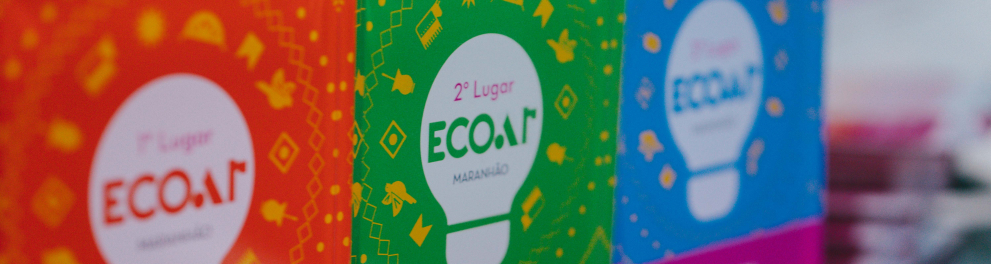 A imagem mostra troféus do prêmio ECOAR Maranhão, organizados lado a lado sobre uma mesa. Em destaque, aparecem premiações de 1º, 2º e 3º lugar, cada uma com cores vibrantes diferentes — vermelho, verde e azul — e o símbolo de uma lâmpada ao centro, associado à ideia de criatividade e inovação.