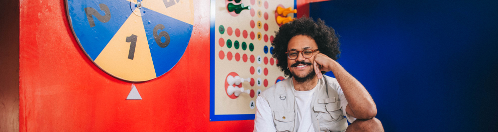 Um homem jovem posa sentado em um pequeno banco de madeira, em um ambiente interno com aparência de estúdio ou espaço expositivo. Ele tem pele morena, cabelo preto volumoso e encaracolado, barba e bigode cheios, além de usar óculos de armação fina. Ao fundo, há um cenário bastante colorido. A parede principal é pintada de vermelho intenso e traz dois painéis de jogos em estilo lúdico.