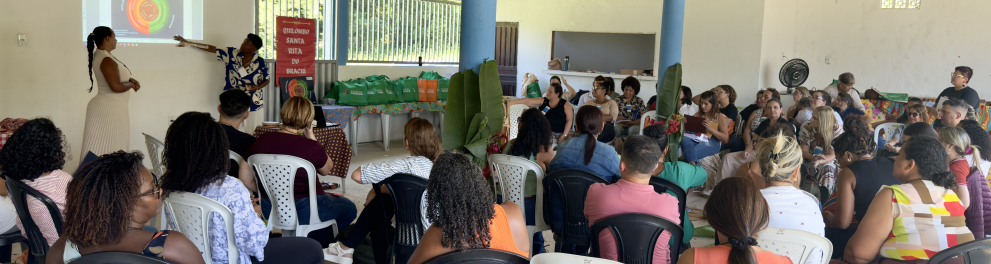 O grupo está reunido em um espaço comunitário, sentado em círculo, acompanhando uma apresentação projetada. O ambiente é simples, aberto e acolhedor, sugerindo uma dinâmica participativa, de troca de conhecimentos e escuta ativa entre educadores ou participantes.