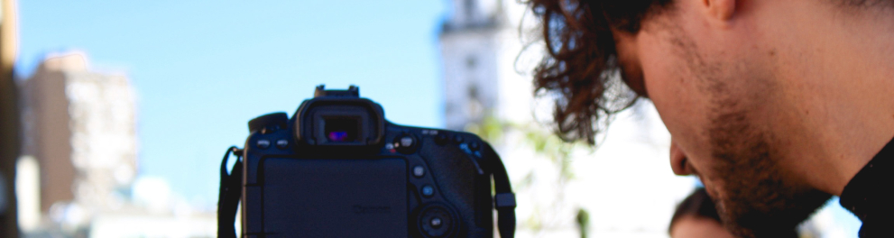 A imagem mostra um jovem operando uma câmera profissional montada em tripé durante uma atividade de gravação ou produção audiovisual. O enquadramento destaca o momento de preparação técnica da filmagem, com foco na câmera e na concentração do operador enquanto ajusta o equipamento.