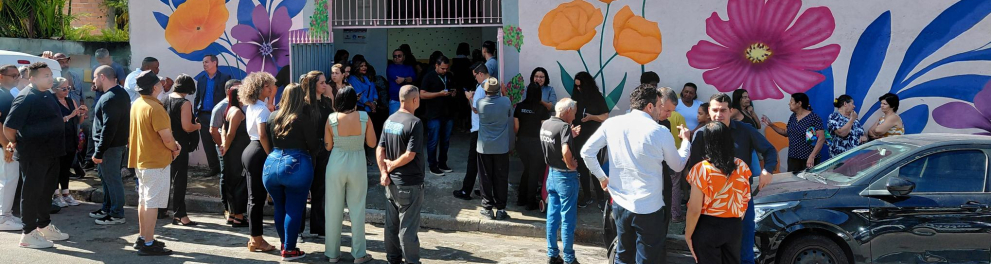 Fachada de um prédio simples com um mural colorido de flores grandes e a placa “CID S.O.S Santa Maria” acima da entrada. Em frente, várias pessoas se reúnem e conversam na calçada, formando pequenos grupos. Um carro preto está estacionado ao lado, e o dia é ensolarado.