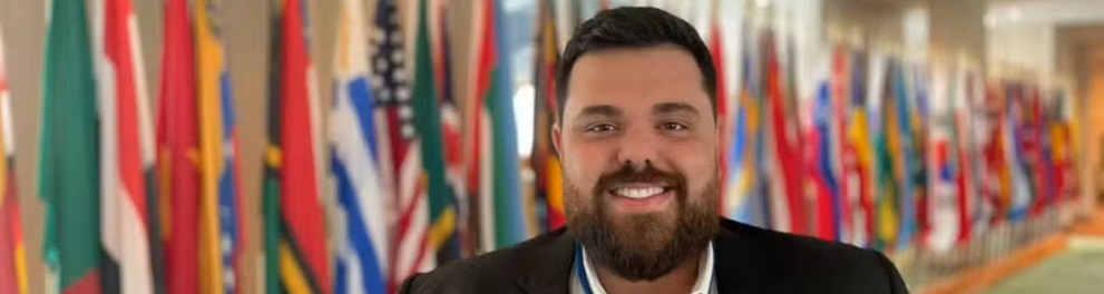 Homem adulto, de barba e cabelo curto escuros, sorri para a câmera usando blazer preto sobre camisa branca e crachá no pescoço. Ele está em um corredor iluminado, com uma fileira de bandeiras de diversos países ao fundo, levemente desfocadas. O ambiente sugere um espaço institucional ou internacional.