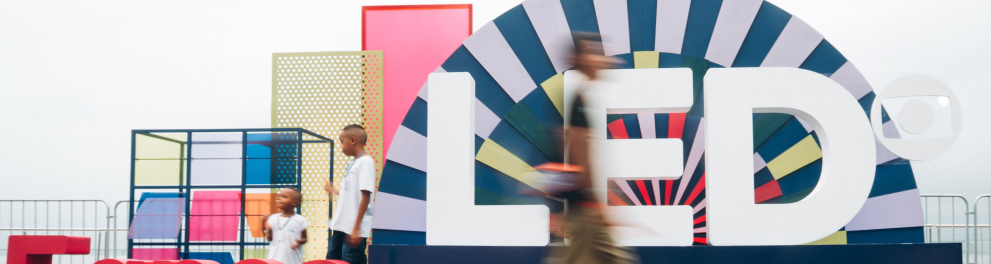 Imagem em ambiente amplo e iluminado, com fundo branco e instalação cenográfica do Festival LED. No centro, há letras gigantes formando a palavra “LED”, em branco, posicionadas diante de um painel semicircular colorido com padrões geométricos em azul, rosa, verde e amarelo. À esquerda, a frase “5 anos” aparece em letras grandes vermelhas sobre um palco azul.