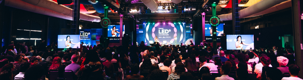 Auditório amplo e escuro, iluminado por luzes coloridas em tons de rosa, azul e roxo. Centenas de pessoas estão sentadas em cadeiras voltadas para um grande palco central, onde acontece uma apresentação do Festival LED – Luz na Educação. No fundo do palco há um telão circular com a identidade visual do evento, cercado por painéis luminosos e dois telões laterais exibindo a palestrante. O teto industrial alto possui estruturas metálicas aparentes e grandes dutos brancos atravessando o espaço