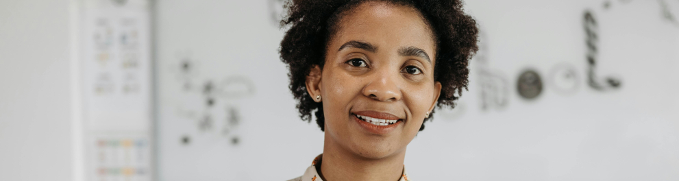 A imagem mostra uma mulher negra de pele clara com um quadro branco ao fundo e segurando um caderno nas maõs fazendo anotações.