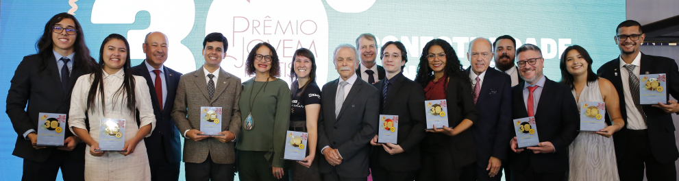 Fotografia de grupo em um palco durante uma cerimônia de premiação. Ao centro, cerca de quinze pessoas, entre jovens estudantes e adultos, posam lado a lado para a foto. Alguns seguram placas ou troféus comemorativos. A maioria está vestida com roupas formais, como ternos, gravatas, vestidos e conjuntos sociais, em cores sóbrias.