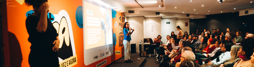 Fotografia de uma palestra em auditório. Em primeiro plano, à esquerda, uma intérprete de Libras aparece em silhueta. No palco, uma mulher com camiseta azul segura um microfone e apresenta conteúdos projetados em uma grande tela. À direita, dezenas de pessoas acompanham atentamente a atividade sentadas em cadeiras organizadas em fileiras. O espaço possui iluminação suave e decoração com a identidade visual do evento em cores vibrantes.