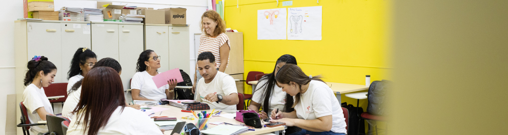 Os estudantes estão sentados em cadeiras, escrevendo, desenhando e conversando, utilizando cadernos, folhas coloridas, canetas, lápis e outros materiais de apoio espalhados sobre a mesa.  Ao centro da cena, uma professora está em pé, sorrindo enquanto acompanha o trabalho da turma. Os participantes usam roupas casuais, a maioria com camisetas brancas, e demonstram concentração e interação durante a atividade.