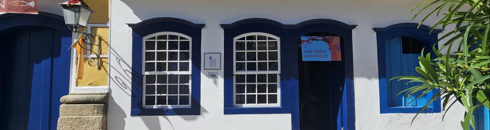 Fotografia da fachada de um casarão histórico localizado em uma rua de pedras. O imóvel tem paredes brancas, janelas e portas em azul-escuro, com detalhes arquitetônicos coloniais, como molduras curvas nas janelas e telhado de cerâmica aparente. No centro da fachada, uma porta aberta dá acesso ao interior da casa, acima da qual há uma placa colorida com a inscrição "casa inspirar para o futuro".