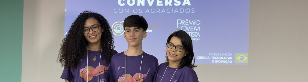 Fotografia de três participantes posando para a câmera em um auditório, em frente a uma tela de projeção. Os três sorriem e estão lado a lado, usando camisetas roxas do Prêmio Jovem Cientista e crachás de identificação pendurados no pescoço.  Ao fundo, a projeção exibe o título "Roda de Conversa com os Agraciados", acompanhado dos logotipos do Prêmio Jovem Cientista, do CNPq e do Ministério da Ciência, Tecnologia e Inovação.