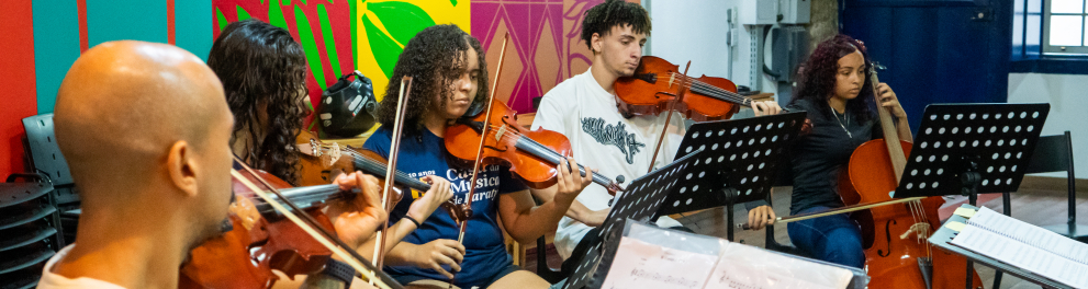 Fotografia de um ensaio musical realizado em ambiente interno. Um grupo de jovens músicos está sentado em semicírculo, concentrado na execução de uma peça para instrumentos de cordas. Em primeiro plano, um violinista aparece de costas para a câmera, enquanto os demais participantes tocam violinos e violoncelos, acompanhando partituras apoiadas em estantes.