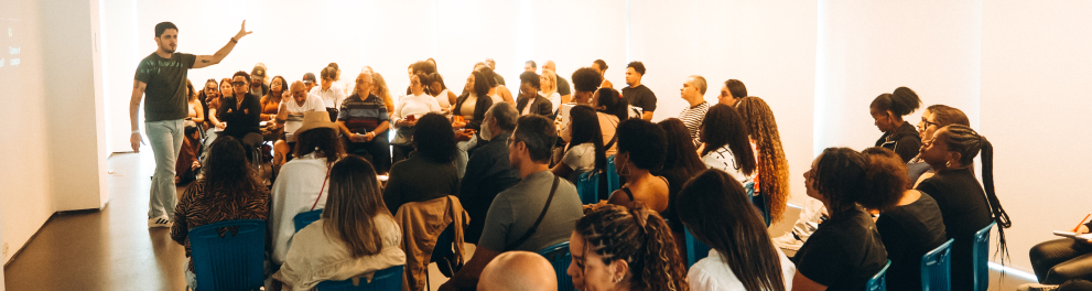 A imagem mostra uma palestra ou atividade de formação realizada em uma sala ampla e iluminada. À frente do ambiente, uma pessoa em pé conduz a apresentação, gesticulando enquanto fala para um público numeroso. Os participantes estão sentados em cadeiras organizadas em fileiras, atentos à exposição; alguns fazem anotações, outros acompanham a atividade ou utilizam o celular.  O espaço possui iluminação artificial, paredes claras e um telão de projeção.