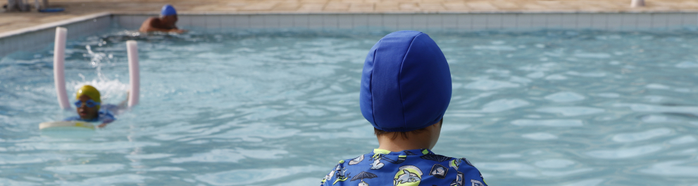 Fotografia de uma piscina durante uma atividade de adaptação ao meio aquático. Em primeiro plano, uma criança aparece sentada de costas na borda da piscina, usando touca de natação azul e camiseta de proteção solar estampada em tons de azul e verde. Ela está apoiada sobre uma prancha de espuma azul e segura um macarrão flutuador curvado ao redor do corpo, observando a atividade na água.
