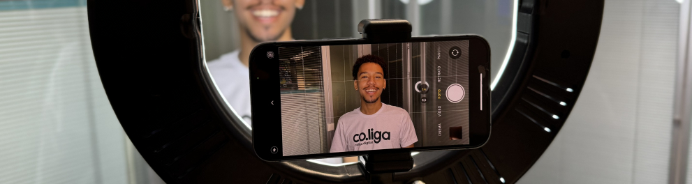Um jovem aparece sendo fotografado ou gravado em um ambiente interno, diante de um grande ring light. Um celular está posicionado no centro do equipamento e enquadra o jovem, que sorri para a câmera. Na tela do aparelho, é possível ver a imagem dele usando uma camiseta branca com a marca co.liga. Ao fundo, há portas e divisórias de vidro, criando um ambiente com aparência de escritório ou estúdio.