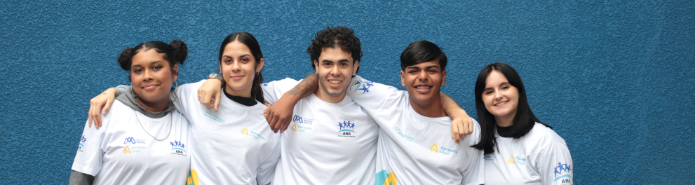 Cinco jovens participantes do Aprendiz Legal posam juntos para uma fotografia. Eles vestem camisetas do programa e aparecem abraçados diante de uma parede azul.
