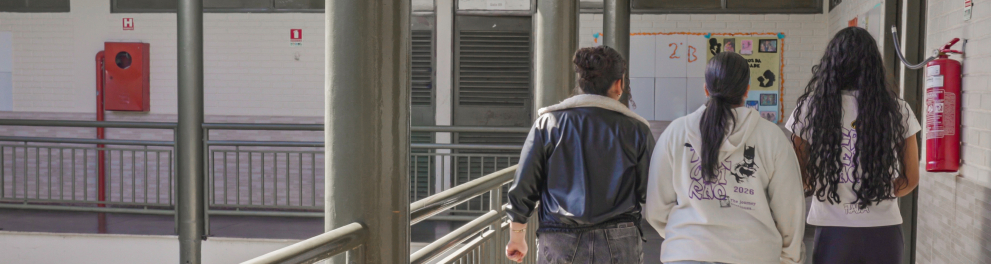 três estudantes caminham por um corredor interno de uma escola, em direção a uma área de salas de aula. O espaço possui corrimãos metálicos, pilares, janelas e murais com trabalhos e produções estudantis. As jovens vestem roupas casuais, e duas delas usam peças com referências a um projeto ou atividade de 2026. A iluminação natural cria sombras marcadas no piso, destacando a arquitetura aberta do corredor.