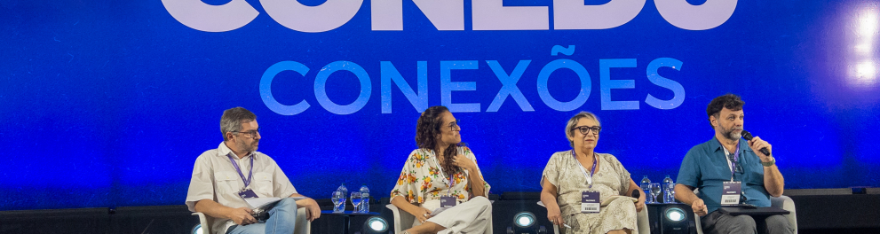 Quatro pessoas sentadas em cadeiras brancas em um palco, com um painel azul ao fundo onde se lê "Palco CONEDU CONEXÕES". Da esquerda para a direita: um homem de camisa bege clara e calça jeans; uma mulher de blusa florida e calça clara; uma mulher de vestido estampado claro e óculos; e um homem de camisa azul, segurando um microfone e falando.