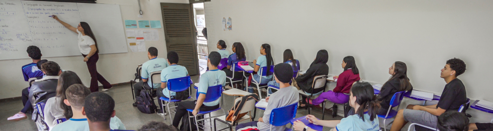 Vista geral de uma sala de aula ampla. À esquerda, uma professora em pé escreve em um quadro branco. Dezenas de estudantes de idades variadas aparecem assentados em carteiras azuis individuais, atentos à explicação, vestindo uniformes escolares em tons de azul e branco ou roupas casuais.
