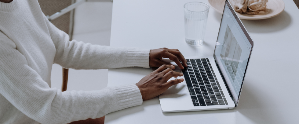 mãos de uma pessoa negra de casaco branco apoiadas na mesa digitando em um laptop