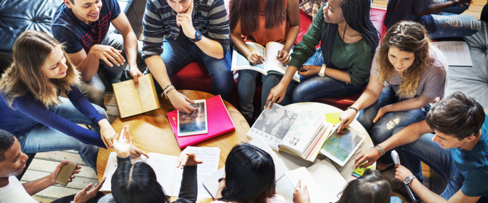 Várias e vários jovens estão sentados ao redor de uma mesa com alguns livros.