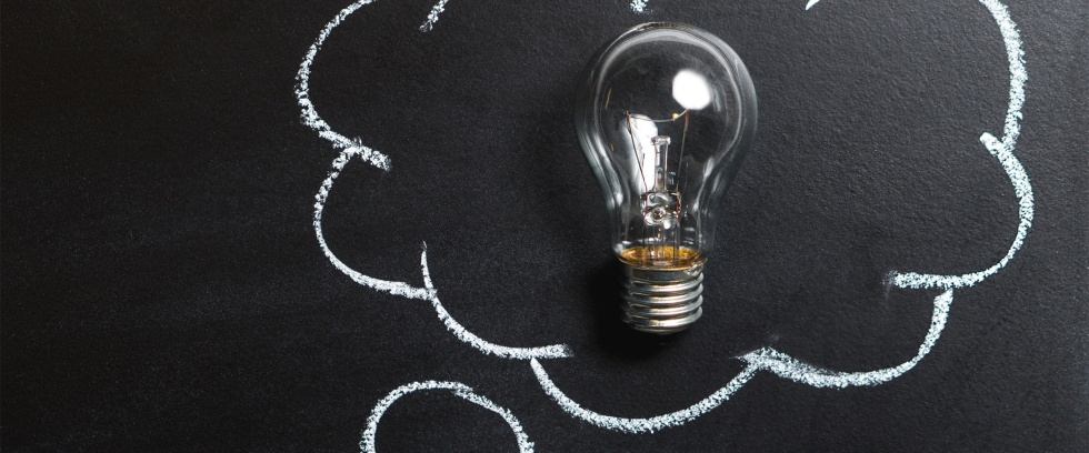 Uma lâmpada está sobre uma superfície escura. Ao seu redor, uma nuvem desenhada com giz sugere que alguém teve uma ideia.
