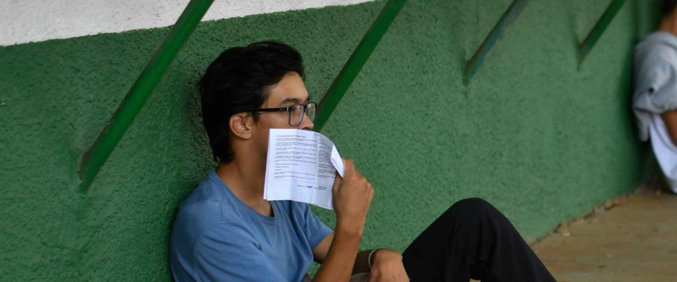 Dois jovens estão sentados no chão, encostas na parede. Ambos estão de calça, tênis e camisa. O que está em primeiro plano tem uma folha de papel na mão e a folha está encostando em seu rosto.