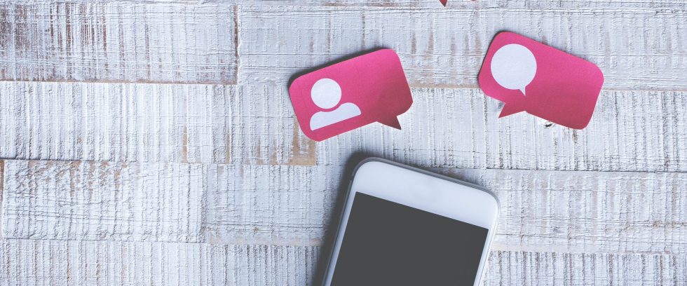 A imagem é uma ilustração. Metade de um celular aparece na parte inferior da imagem. Próximas a ele, caixas de diálogo rosa com corações brancos.