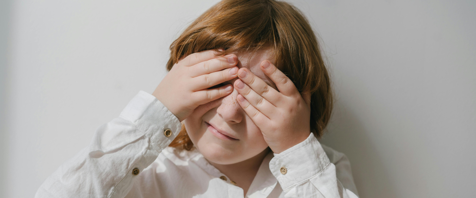 Uma criança branca de cabelos lisos e castanhos está tapando os dois olhos com as suas próprias mãos