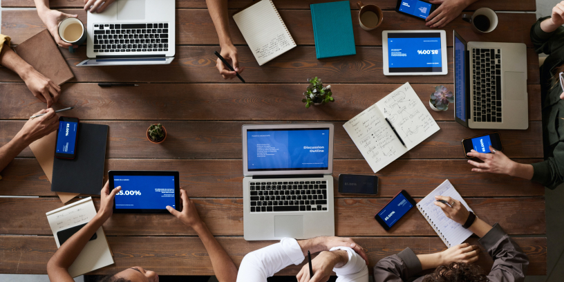 Diversos computadores dispostos sobre uma mesa com algumas pessoas sentadas ao redor dela.