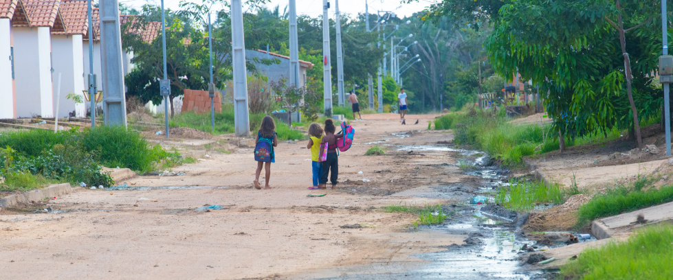 Ao longe, as crianças vão a pé para a escola em uma rua não asfaltada e com esgotão a céu aberto