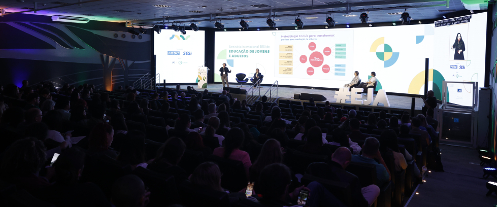 Auditório moderno com cadeiras ocupadas por diversas pessoas assistindo a uma apresentação. No palco iluminado, há quatro pessoas — duas em pé e duas sentadas — participando de um painel com telão ao fundo. No telão está escrito: “Seminário Internacional SESI de Educação de Jovens e Adultos”, com gráficos e textos explicativos. À direita, uma tela vertical mostra uma intérprete de Libras (Língua Brasileira de Sinais). O ambiente tem iluminação colorida nas laterais e teto com painéis de LED.