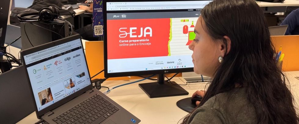 Uma mulher de cabelos longos e escuros, usando brinco de prata, está sentada em frente a duas telas em um ambiente de escritório. Ela olha para um monitor onde está aberto o site do curso “SEJA – Curso preparatório online para o Encceja”. À sua esquerda, há um notebook com a página inicial da Plataforma Futura aberta, exibindo programas como Co.liga, Telê Curso e Aprendê Legal. No fundo, outras pessoas também estão estudando. O ambiente é moderno, com iluminação clara e clima de concentração.