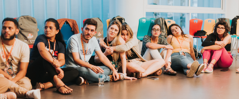 Um grupo de jovens está reunido em roda dentro de uma sala ampla, iluminada por luz natural. Todos estão sentados no chão ou em cadeiras, em clima de descontração e escuta. Ao centro, uma jovem fala enquanto os demais observam com atenção. O ambiente é acolhedor e transmite o sentimento de encerramento e afeto entre o grupo.