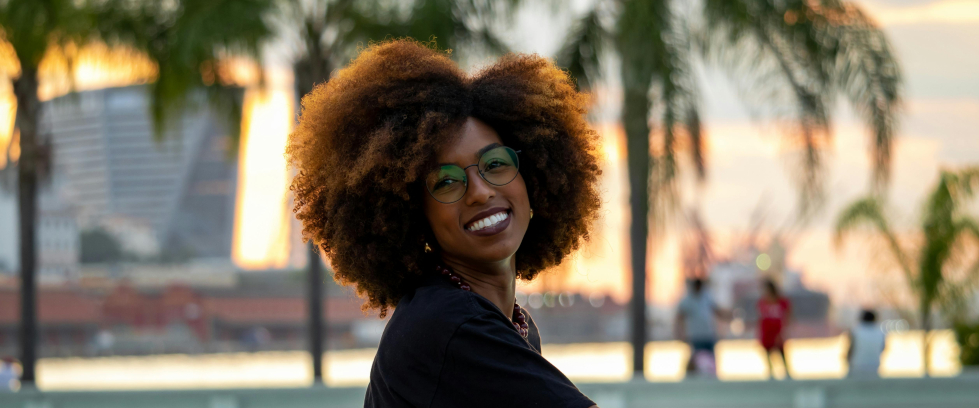 Uma mulher negra sorrindo e sentada e posando de lado para uma foto em frente ao espelho d'água do Museu do Amanhã