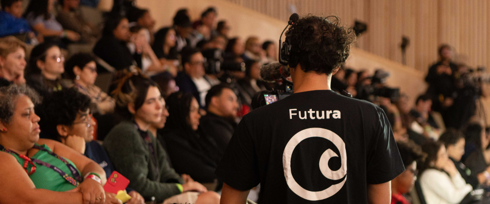 Plateia de um auditório lotado, com pessoas sentadas e atentas à programação. Em primeiro plano, de costas, está um cinegrafista usando fones de ouvido e camiseta preta com o logotipo do Canal Futura, que grava o evento. Algumas pessoas do público seguram celulares e objetos coloridos nas mãos. O ambiente tem iluminação suave e paredes revestidas de madeira clara.