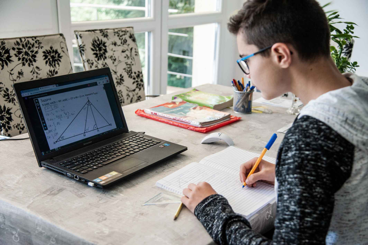 Um jovem, que usa óculos, está escrevendo algo em uma folha branca enquanto observa o que está na tela do laptop.