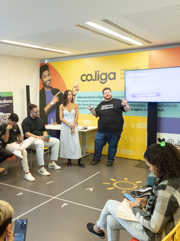 Cerca de 20 pessoas participam de uma oficina realizada em uma sala clara e colorida, com paredes decoradas com as marcas “co.liga” e “co.educa”. A maioria dos participantes está sentada em cadeiras amarelas e vermelhas, organizadas em semicírculo, com atenção voltada para dois facilitadores em pé ao centro — uma mulher de vestido claro e um homem com camiseta preta onde se lê “Transformando realidades”. Muitos usam celulares ou fazem anotações. À direita, uma tela exibe a frase “E pra começar...”, com inst