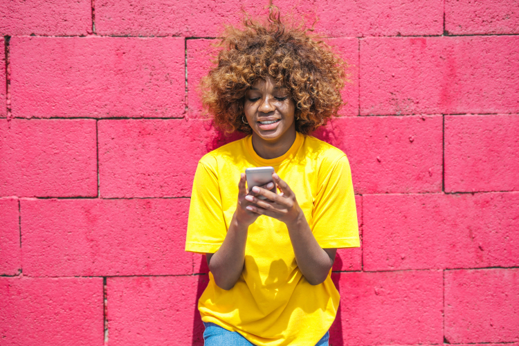 uma jovem de camisa amarela com um celular nas mãos e uma parede de tijolos rosa ao fundo