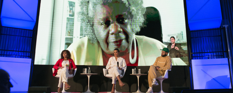 Três pessoas estão sentadas em poltronas. Ao fundo, a imagem de Conceição Evaristo, uma mulher negra, com cabelo crespo e grisalho, é projetada em tamanho maior.