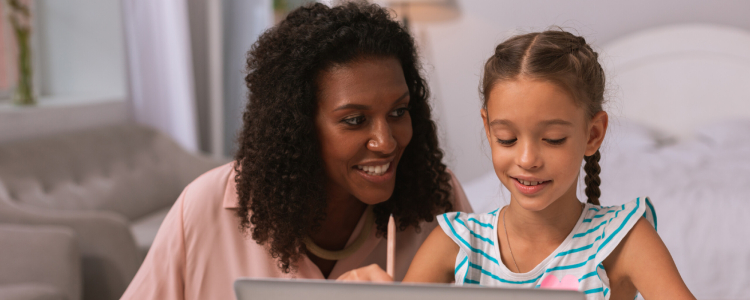 Uma mulher negra auxilia, sorrindo, uma criança a escrever. A criança também está sorrindo.