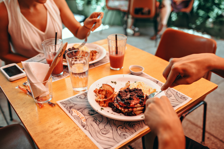 Duas pessoas estão diante de seus pratos de comida sobre a mesa de um restaurante a céu aberto. Seus rostos não estão enquadrados na foto.